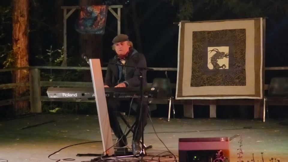Viktor at a keyboard on an outdoor stage beside a framed artwork.