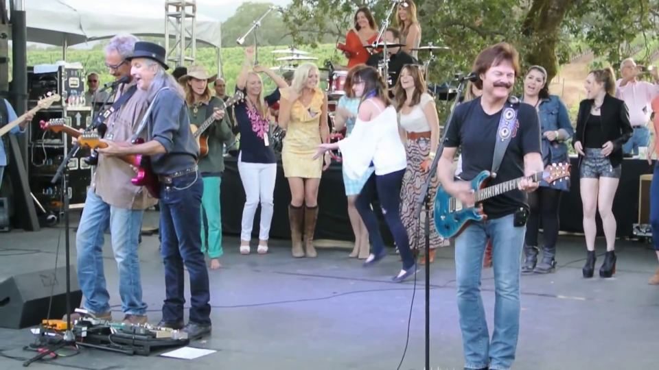 The Doobie Brothers on an outdoor stage at BR Cohn Winery, guitars up, with a crowd dancing behind them.