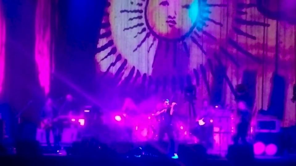 A stage lit in purple and pink with a large sun banner behind the band.