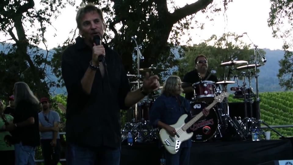 Kenny Loggins singing at the microphone with the band behind him under the trees.